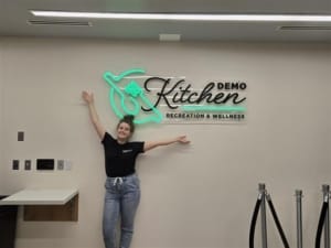 UVU Student Olivia Ellis next to the Student Demo Kitchen logo in the Student Life and Wellness Center during an event | Photo by Kenna Seegmiller